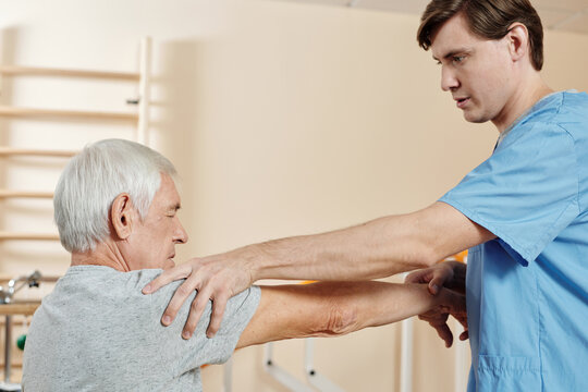Professional male nursing assistant exercising hand of senior Caucasian man getting through physical therapy course in rehab center