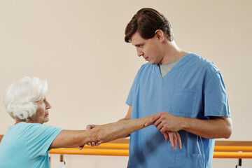 Professional male physiotherapist checking and exercising hand of senior Caucasian woman getting physical therapy in rehabilitation hospital