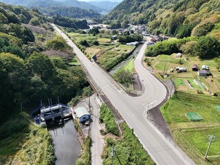 A road with a bridge over a river and a small town in the background. The road is wide and straight, with a few trees in the distance