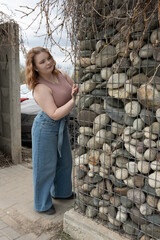 Fototapeta premium Young lady standing beside a wire mesh gabion with rocks. Modern landscape design element.