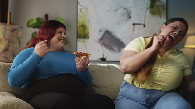 Two cheerful curvy women singing with imaginary mic and eating pizza at home