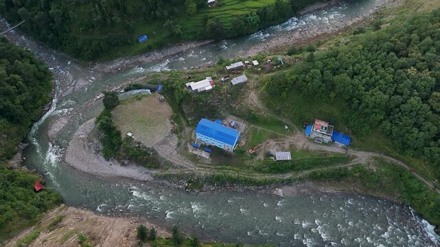 Aerial perspective looking down at a mountain river confluence and a remote rural facility with bright blue roofs in a green valley