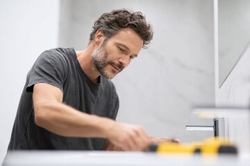 Mature man focused on DIY home improvement project in a bright, modern bathroom. Repair, maintenance, or renovation concept. Authentic lifestyle image.