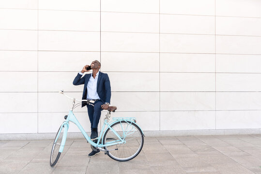 Happy businessman talking on smartphone while standing with his bicycle near white wall
