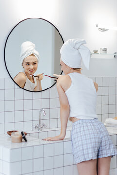 Blonde with a white towel on her head brushes her teeth in front of a mirror in a white bathroom.