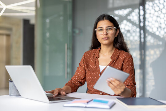 Indian businesswoman at modern office desk using laptop and tablet, focused on multitasking and productivity while planning and communicating in a professional workplace setting - Powered by Adobe