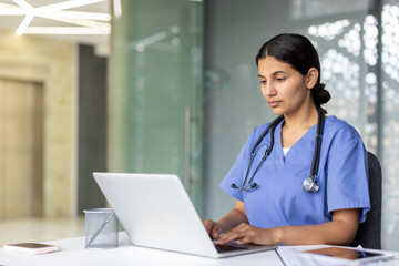 Dedicated healthcare professional wearing scrubs and a stethoscope, working on a laptop at a desk within a modern medical facility, representing telemedicine and digital healthcare