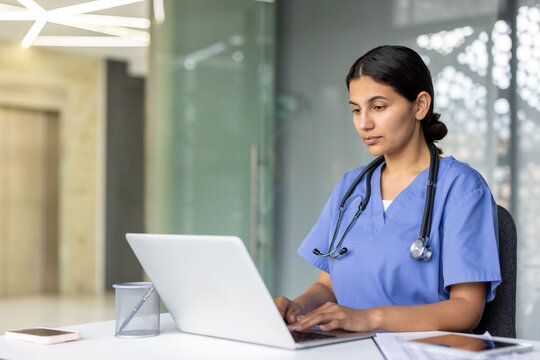 Dedicated healthcare professional wearing scrubs and a stethoscope, working on a laptop at a desk within a modern medical facility, representing telemedicine and digital healthcare