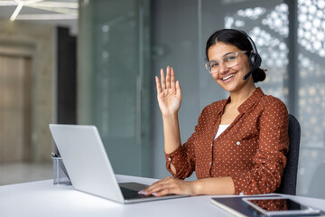 Friendly professional woman in glasses wearing a headset, smiling and waving during a video call from a modern office desk with laptop, demonstrating remote support and communication
