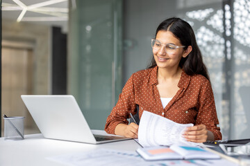 Indian businesswoman smiling, wearing glasses, analyzing financial documents and using a laptop while sitting at a desk in a contemporary office setting