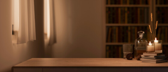 Alcohol bottle and glass cup with candles over book on wooden table across bookshelf in dim lit room