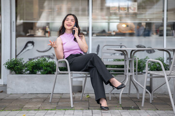 Young asian woman holding and talking on phone with a smile while sitting at table in front of cafe.