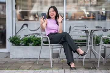Young asian woman talking video call on phone with a smile while sitting at table in front of a cafe
