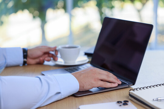 Man with cup of drink working on laptop at wooden table indoors, closeup - Powered by Adobe