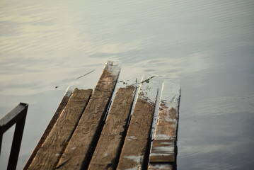Fototapeta premium An old wooden pier. An old wooden walkway stands on rusty iron supports in the water. The wood has turned black with age. The ends of the planks have sunk into the water. Sky is reflected from water.