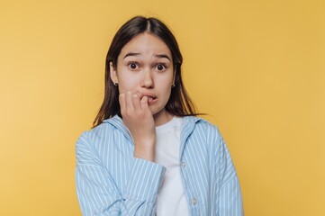 Obraz premium Young woman biting nails, looking anxious against yellow background.