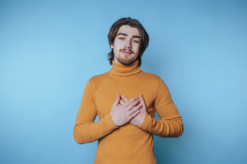 Young man in mustard turtleneck with hands on chest against blue background.