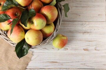 Fresh ripe apples with green leaves in wicker basket on white wooden table, top view. Space for text