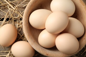Raw chicken eggs in bowl and straw on wooden table, flat lay