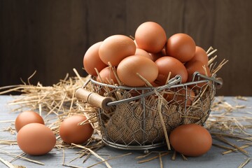 Raw chicken eggs, metal basket and straw on grey wooden table against brown background, closeup