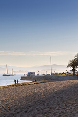 A scenic view of a tranquil beach in Santander, Spain, with sailboats on the water and mountains in...