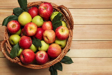 Fresh apples in wicker basket and green leaves on wooden table, top view. Space for text
