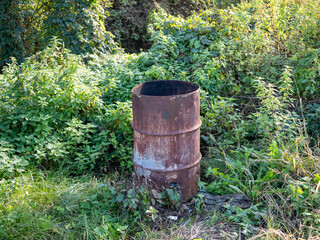 Obraz premium Old rusty metal barrel used for burning waste, standing outdoors surrounded by green vegetation and grass.