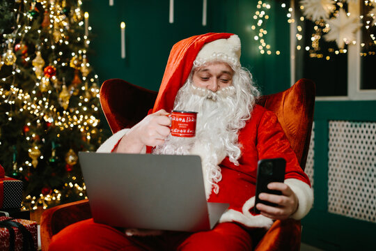 Santa claus using laptop and smartphone, drinking from christmas mug