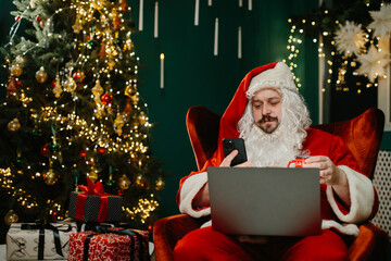 Santa claus using laptop and smartphone near christmas tree