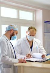 Two male doctors in protective medical masks discussing patient diagnosis and reviewing medical documents in a hospital. Concept of healthcare teamwork, professional medicine, and hospital routine.
