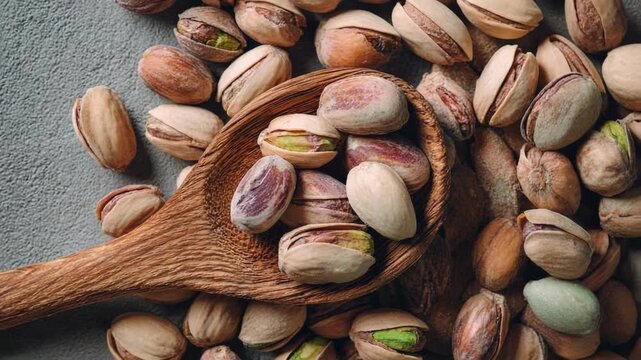 Pistachio nuts in a wooden spoon with shells scattered around on a gray surface.
