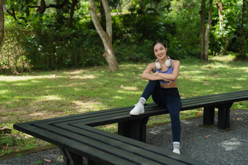 Fototapeta premium Young Asian woman in sportswear sits on a park bench with wireless headphones around her neck, smiling at the camera while enjoying a restful moment outdoors after a workout