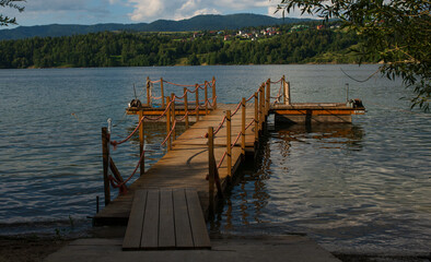 Fototapeta premium Wooden pier with orange ropes stretches into a quiet lake surrounded by forested hills and small village houses. Evening sunlight creates reflections and a sense of calm countryside beauty.