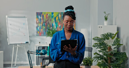 A beautiful and confident businesswoman stands in an open space office, holding a tablet, working on reports, responding to emails, and smiling at the camera, focused on her tasks.