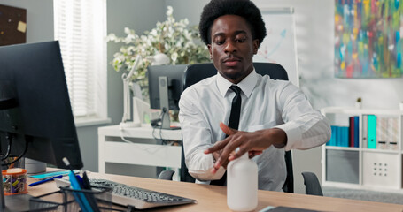 The company boss in a white shirt and tie sits at his desk, squeezing disinfectant from a dispenser, sanitizing his hands before touching the laptop keyboard, ensuring office hygiene and safety.