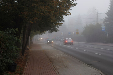 Obraz premium Car driving through foggy morning streets in the town, low visibility and difficult road conditions