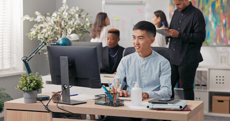 An Asian man sits in front of an office computer, using a web browser to manage the company's social media. He works on the screen, creating ads, posts, and graphics for online marketing.