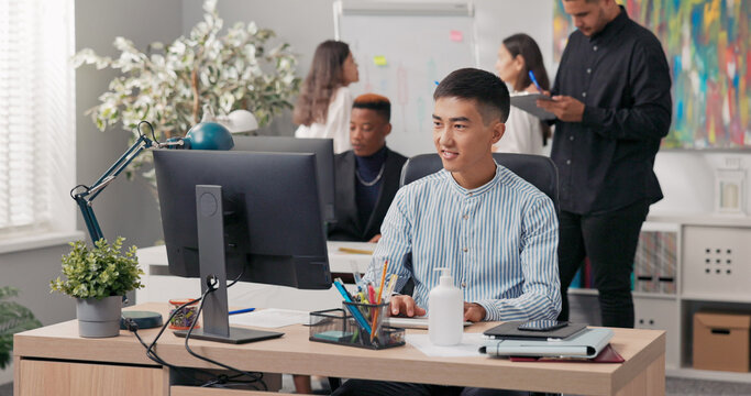 An Asian man sits in front of an office computer, using a web browser to manage the company's social media. He works on the screen, creating ads, posts, and graphics for online marketing.