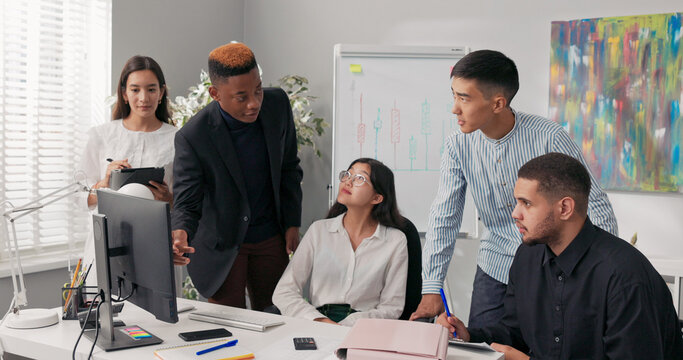 A group of company employees work on a project, sitting at a computer. A man in a suit holds sticky notes in his hand, sticking them on the monitor as they plan and organize the project. - Powered by Adobe