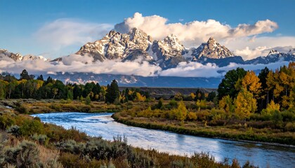 Majestic mountain range rises behind a winding river with vibrant autumn foliage on its banks under a bright blue sky dotted with clouds