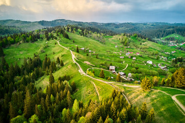 Aerial photograph of serene rural landscape in Ukraine, featuring rolling green hills, winding dirt roads, and scattered houses. Dense forests surround area, with dramatic, cloudy sky overhead.