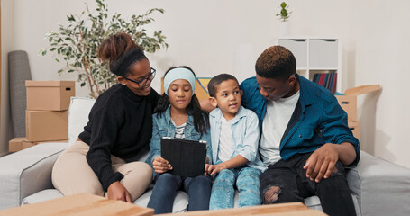The family spends time together on the couch, the parents with their two children browse the furniture for the new apartment on website on tablet, the siblings choose decorations for room