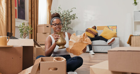 A woman sits in the middle of her new home surrounded by boxes. She finds a vase in one of them,...