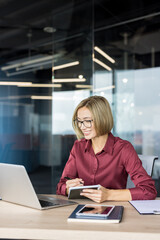 Obraz premium Smiling businesswoman in glasses taking notes while working on a laptop at her desk in a contemporary office, representing productivity and professional success