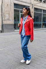 A teenager in a red jacket and blue jeans stands on a sidewalk