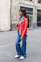 A teenager in a red jacket and blue jeans stands on a sidewalk