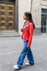 A teenager in a red jacket and blue jeans stands on a sidewalk