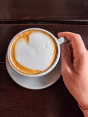 Hand holding a cup of cappuccino with heart-shaped foam on a saucer in a cozy cafe setting