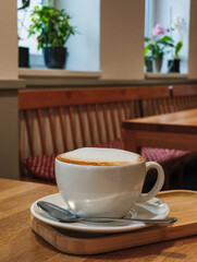 Coffee cup with frothy milk on wooden tray in cozy cafe setting with plants and warm ambiance