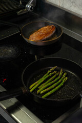 Two frying pans on an electric stove, one with green asparagus and the other with a duck breast, for a healthy gourmet meal.